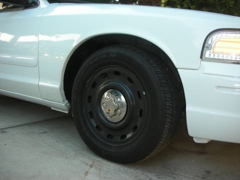 2004 Ford Crown Victoria - Close-up of the passenger-side front wheel on a white Ford Crown Victoria parked on concrete, fitted with a plain black steel wheel, small…