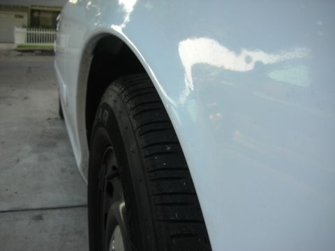 2004 Ford Crown Victoria - Tight low angle along the glossy white rear quarter panel of a sedan parked on a concrete driveway, with the wheel arch framing a Goodyear…