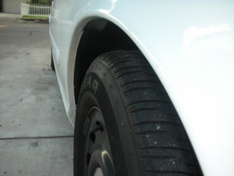 2004 Ford Crown Victoria - Low, tight angle along the side of a white sedan on a concrete driveway, framing the rear wheel arch and a Goodyear tire with fresh tread o…