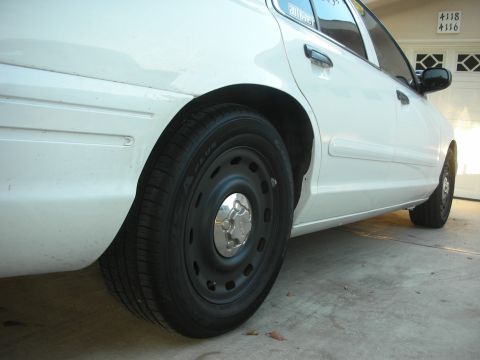 2004 Ford Crown Victoria - Low side view down the flank of a white Ford sedan on a driveway, focused on the front wheel: a black painted steel wheel with a small chro…