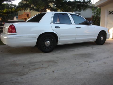 2004 Ford Crown Victoria - A white Ford Crown Victoria sedan viewed from the rear passenger 3/4 angle, parked in a concrete driveway with black steel wheels and a sma…