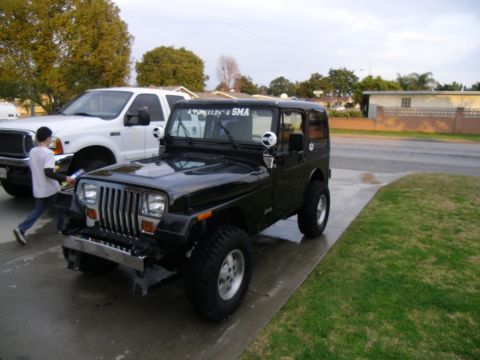 1992 Jeep Wrangler - A black 1992 Jeep Wrangler is parked next to a white pickup truck. The Jeep has a custom bumper and text on the windshield.
