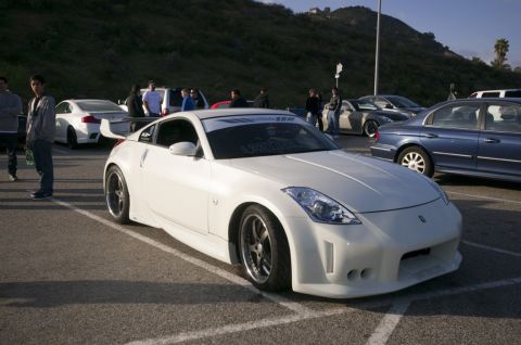 2006 Cadillac STS - A white Nissan 350Z is parked in a lot with other vehicles and people around.