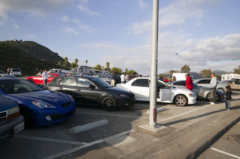 Cars parked in a lot with people standing around, RVs in the background