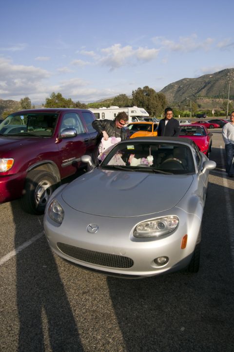 2006 Cadillac STS - A silver Mazda MX-5 Miata convertible is parked next to a red truck, with other vehicles and people in the background.