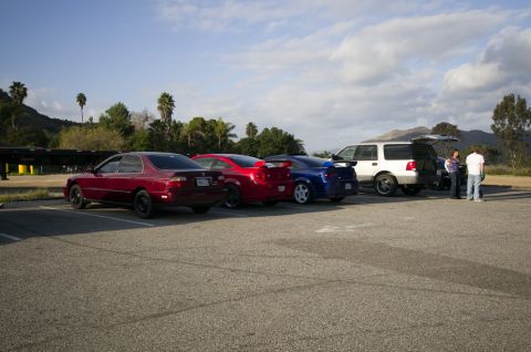 2006 Cadillac STS - A lineup of cars and an SUV are parked in a lot under a partly cloudy sky with mountains visible in the distance.