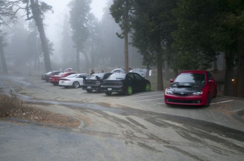 2006 Cadillac STS - A red Subaru Impreza WRX with a black hood is parked in a lot with other cars on a foggy day.