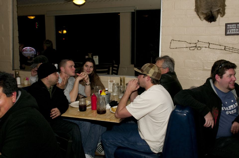 People gathered around tables in a dimly lit restaurant, with a rifle decoration on the wall