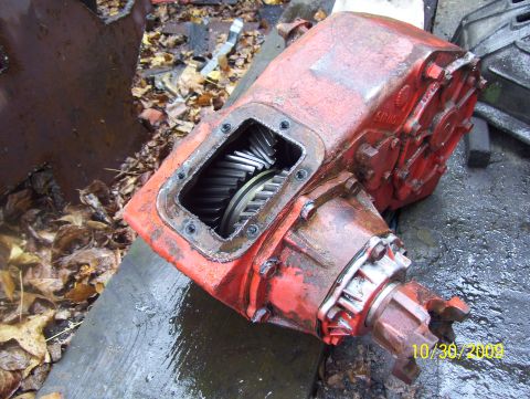 1978 Ford Bronco - This red transfer case, with its gears exposed, rests on a wet surface amidst fallen leaves.