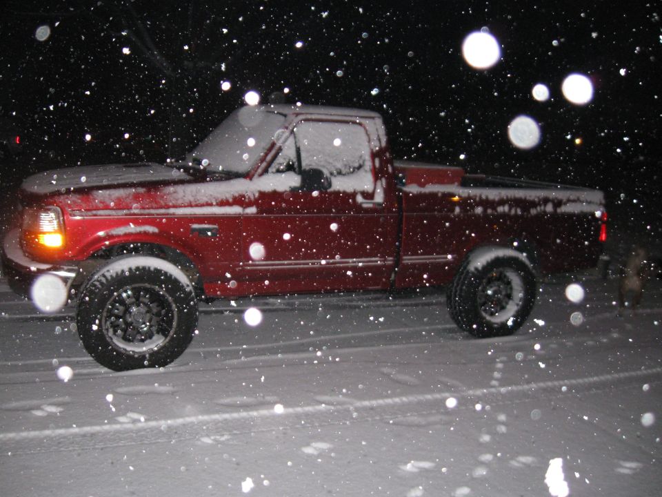 Red pickup truck driving in snow at night. It has a single cab and a short bed