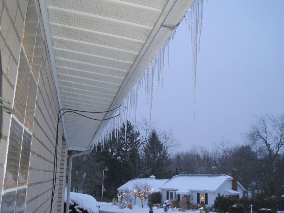 Icicles hang from the white soffit of a house, with snow-covered yards and trees visible