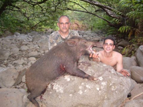 1991 Ford Bronco - Two men crouch in a boulder-strewn jungle streambed behind a large tusked wild boar draped over a rock, one of them holding up the boar's t…