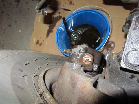 1984 Ford Bronco - Close-up of a vehicle's brake caliper and tire, with a blue bucket containing dark fluid in the background.