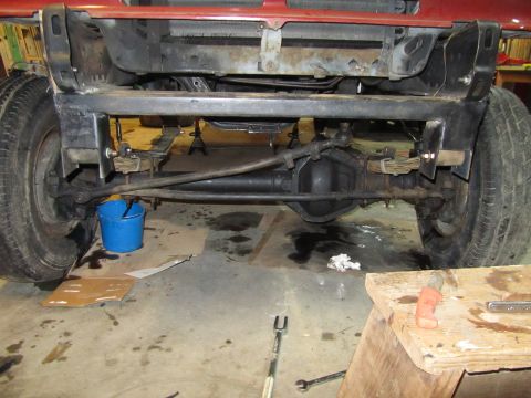 1984 Ford Bronco - Underneath view of a vehicle's front axle and suspension components, with tools and a bucket nearby.