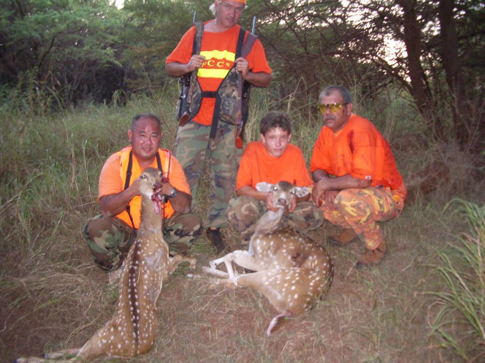 Hunters in blaze orange and camo posing with two axis deer in tall grass