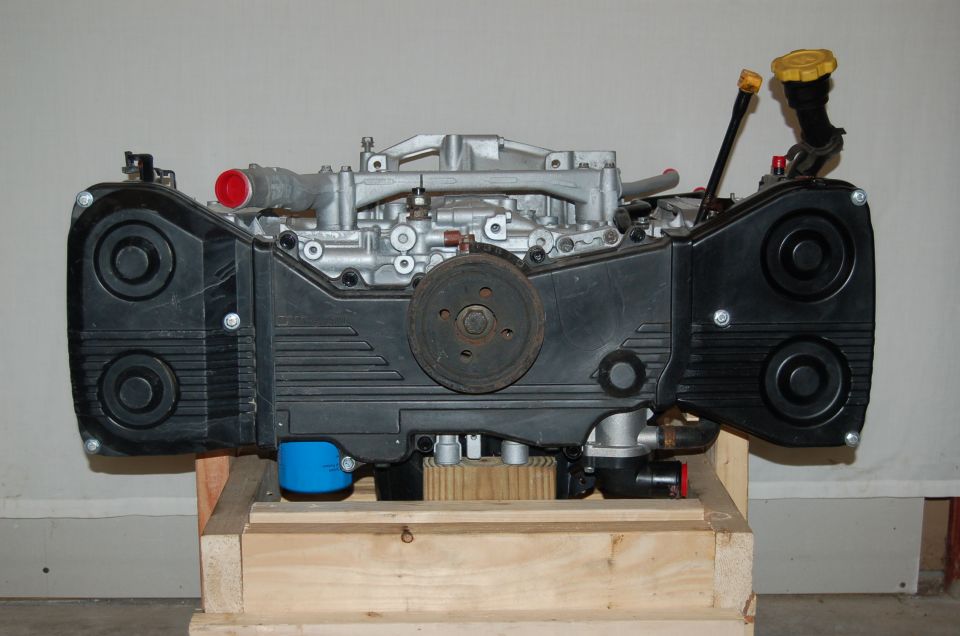 Subaru boxer engine on a wooden crate, front view with black timing covers and yellow oil cap