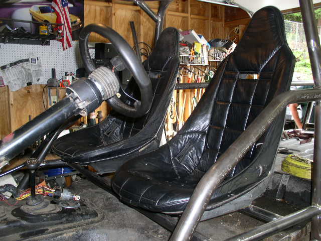 Interior view of a vehicle with two black racing seats, a steering column, and a roll cage. A yellow bag is visible on the right