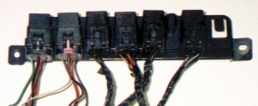 1990 Ford Bronco - Six black cube-style automotive relays plugged into a shared metal mounting bracket with slotted bolt holes at each end. Wiring pigtails in…