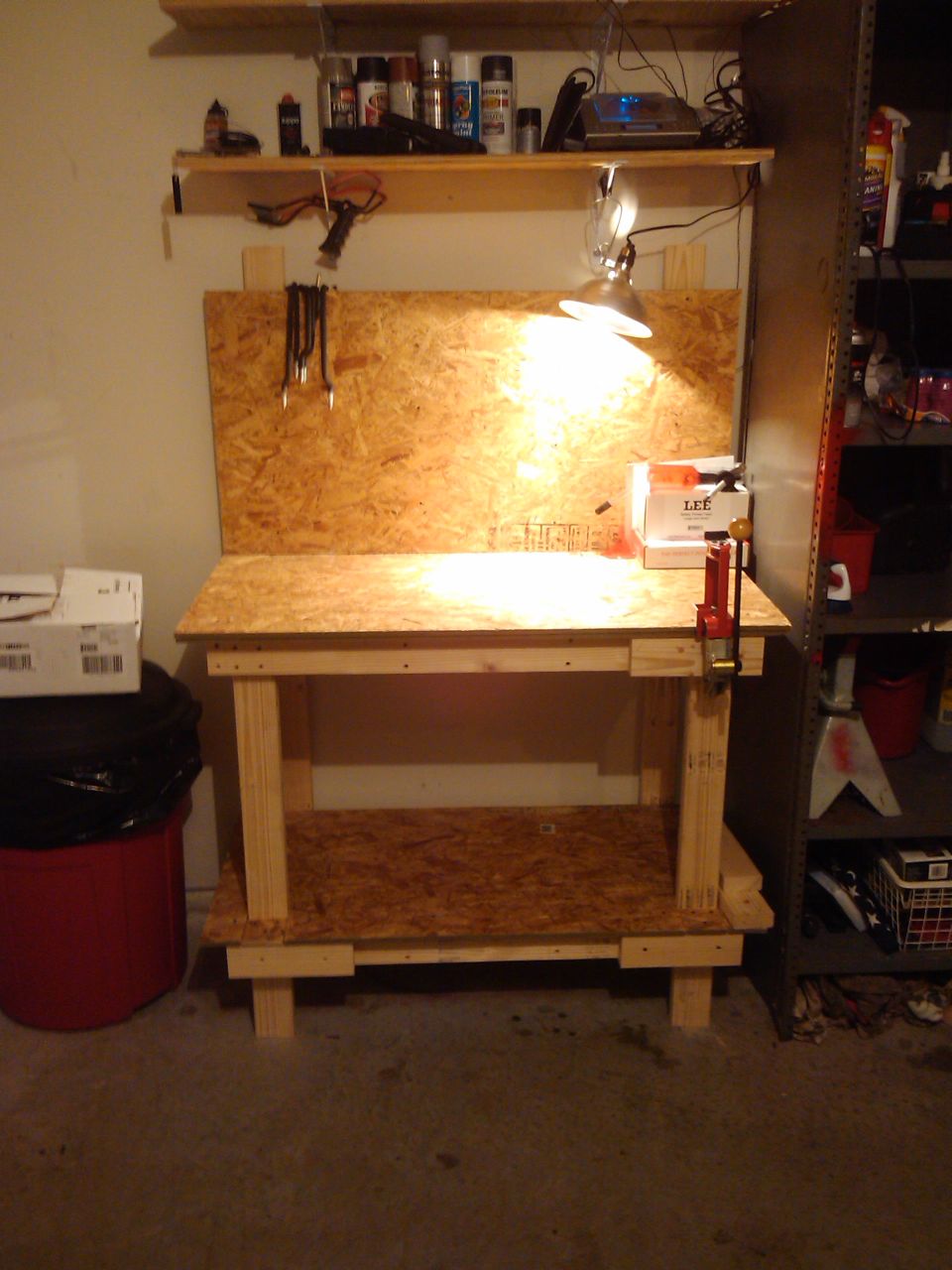 Workbench with shelves above, tools hanging, and a red trash can. A box labeled 'LEE' is on the bench