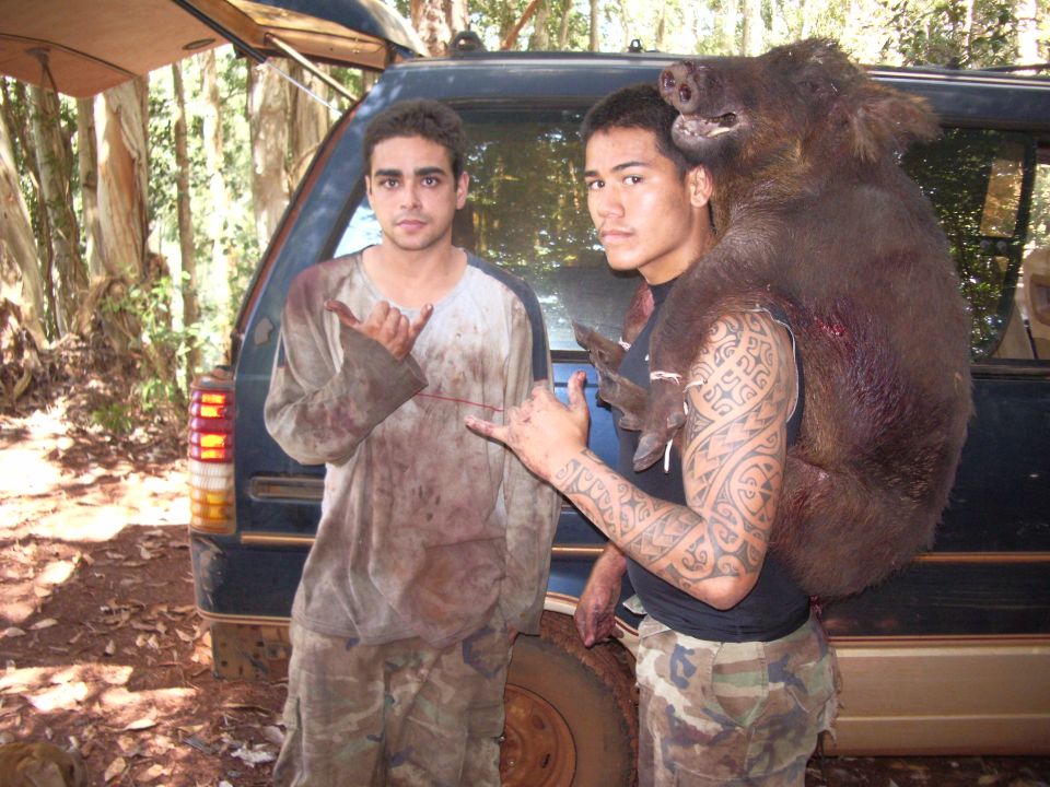 Two guys stand by a dark SUV in the woods, one carrying a large wild boar over his shoulder
