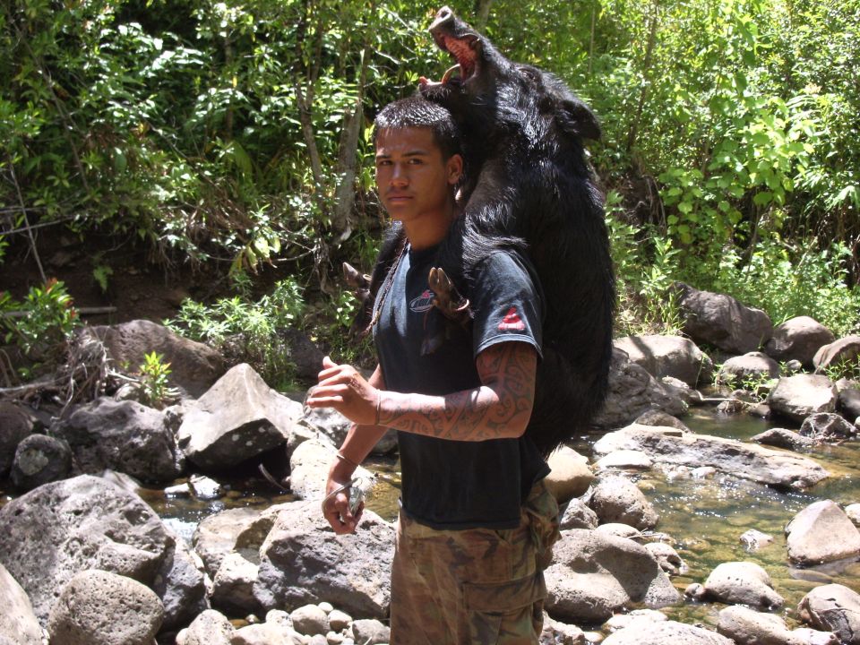 Man with tattoos carries a large wild boar over his shoulders in a rocky, wooded area near a stream