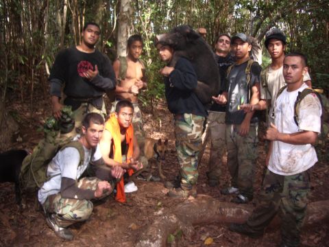 1991 Ford Bronco - A group of nine hunters in camouflage pants and backpacks pose on a forest trail with hunting dogs, one man carrying a large dark boar acro…