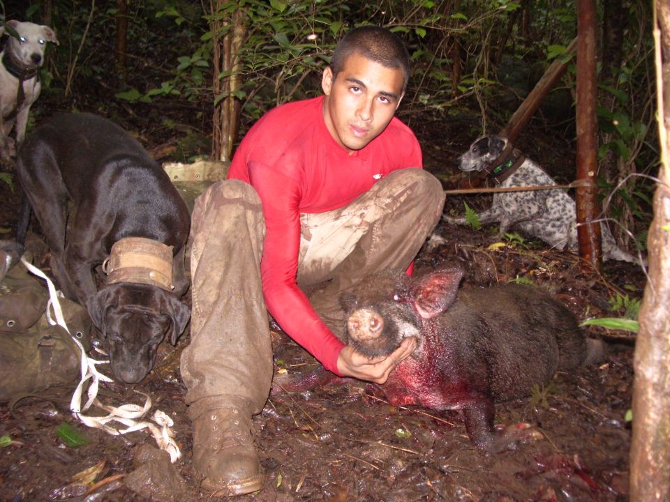 Man in red shirt and muddy pants holds a dead hog, with three dogs nearby in a wooded area