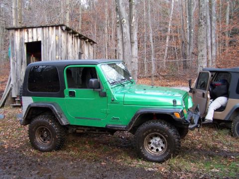 2004 Jeep TJ - Two Jeep Wranglers parked in a muddy, wooded area with a small wooden structure in the background.