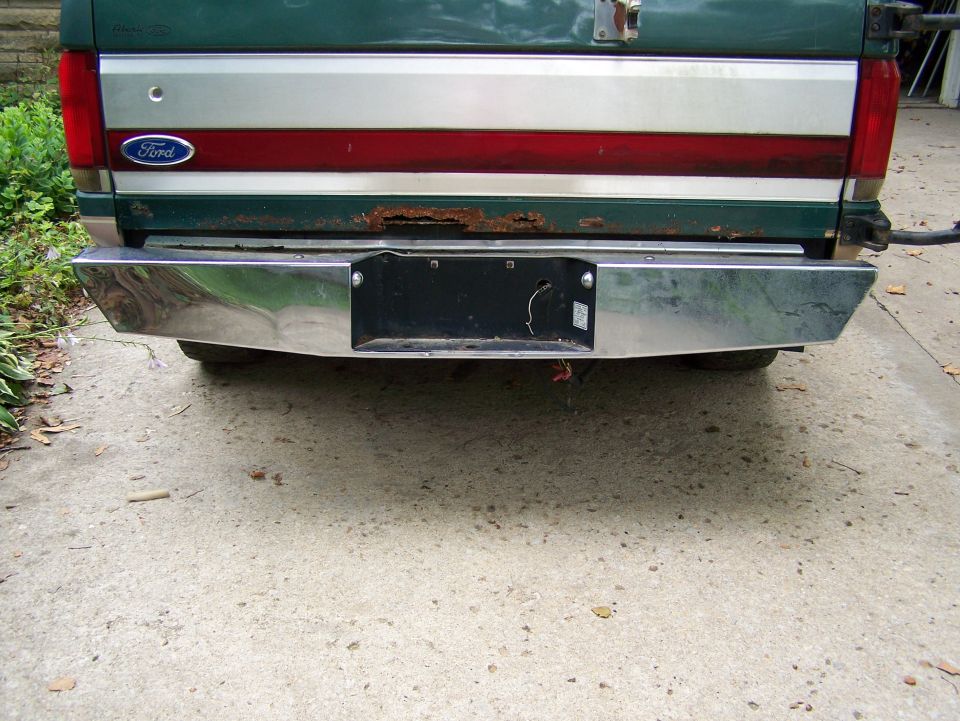 Rear of a green Ford pickup with a chrome bumper and a red stripe on the tailgate. Some rust is visible