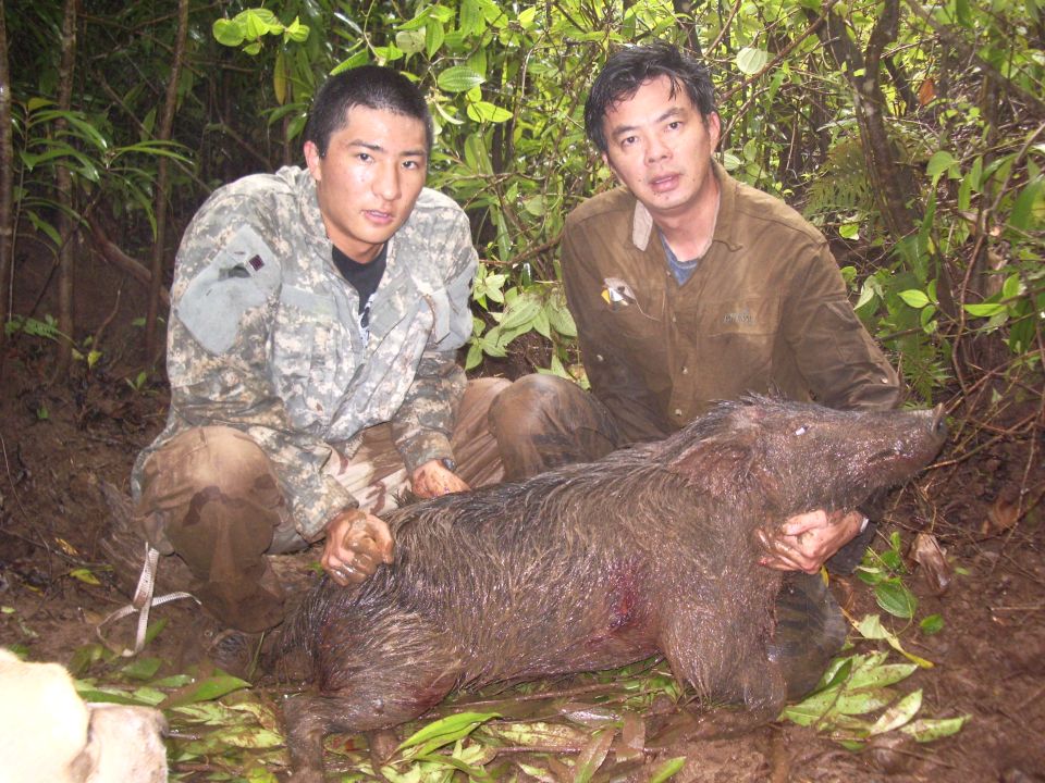 Two men in hunting attire pose with a large wild boar in a wooded area