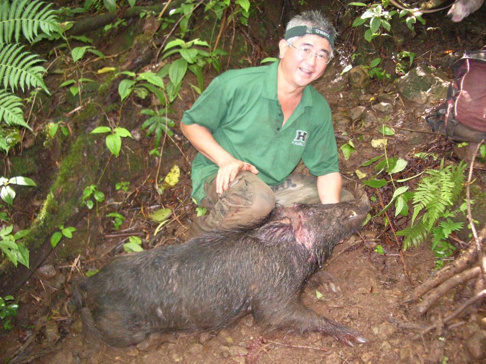 Man in green shirt and camouflage pants kneels next to a wild boar on a muddy forest floor