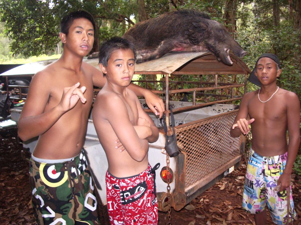 Teens beside a mesh dog-box utility trailer with a boar on its wooden roof