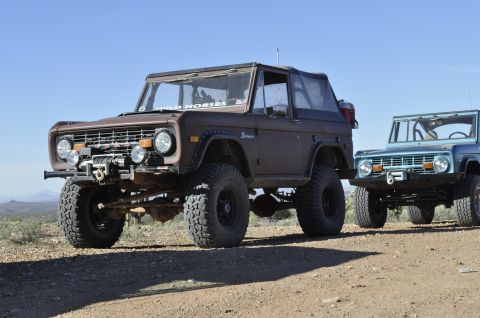 1991 Ford Bronco - Two Ford Broncos, one brown and one blue, are parked on a dirt road under a clear sky.