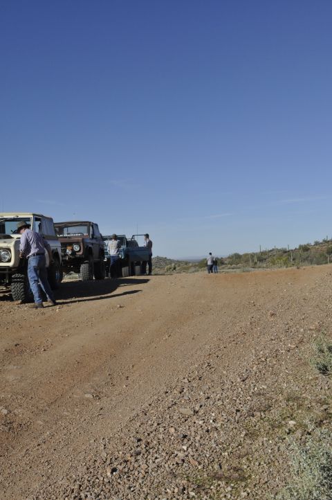 1991 Ford Bronco - Multiple off-road vehicles are parked on a dusty dirt road under a clear blue sky.