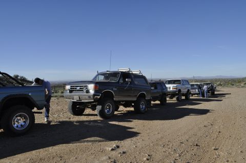 1991 Ford Bronco - A line of SUVs and trucks are parked on a dirt road under a clear blue sky.