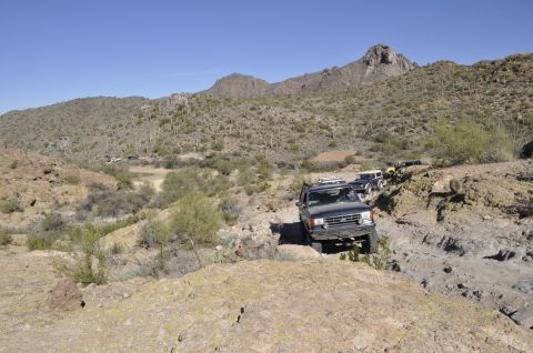 1991 Ford Bronco - Several SUVs navigate a challenging off-road trail through a desert landscape with mountains in the background.