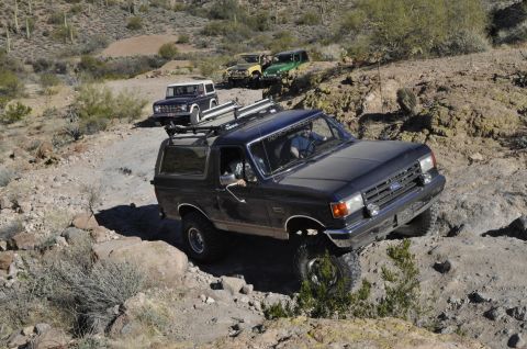 1991 Ford Bronco - Multiple Ford Broncos navigate a challenging off-road trail in a desert landscape.