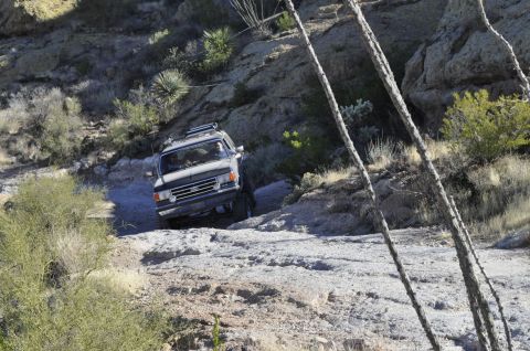 1991 Ford Bronco - A dark SUV with a roof rack is shown off-roading on a steep, rocky trail.