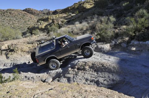 1991 Ford Bronco - A dark gray 1991 Ford Bronco navigates a challenging rocky terrain under a clear blue sky.