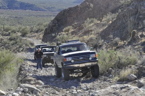 1991 Ford Bronco - Multiple Ford Broncos navigate a challenging rocky trail in a desert landscape.