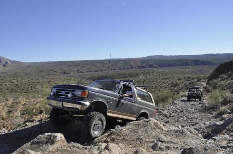 1991 Ford Bronco - A dark gray 1991 Ford Bronco navigates a , rocky desert path under a clear blue sky, with another off-road vehicle following.