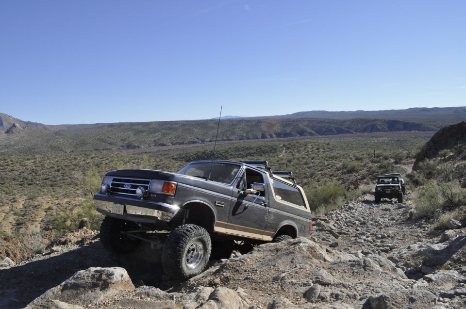 Dark gray 1991 Ford Bronco with tan stripe on rocky desert trail, another vehicle behind
