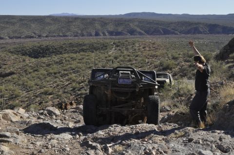 1991 Ford Bronco - A modified off-road vehicle navigates a rocky, desert landscape with a person celebrating nearby.