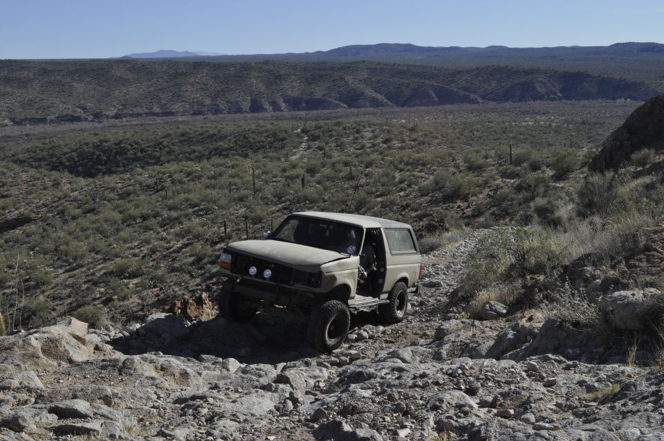 Tan Ford Bronco on rocky terrain, driver's door open, front grille visible