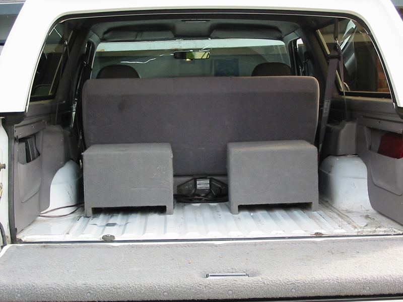 Rear cargo area of a white SUV with gray bench seat and two carpeted speaker enclosures on the ribbed floor