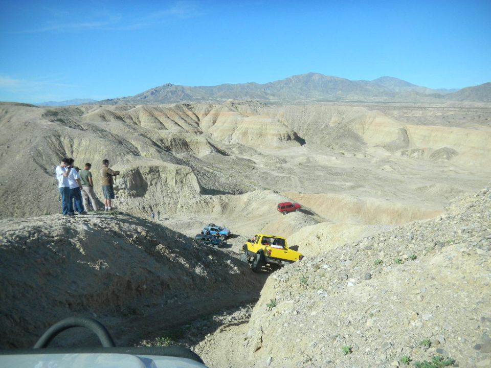 Yellow 1997 Ford Ranger driving down a dirt hill in a desert with other off-road vehicles and people