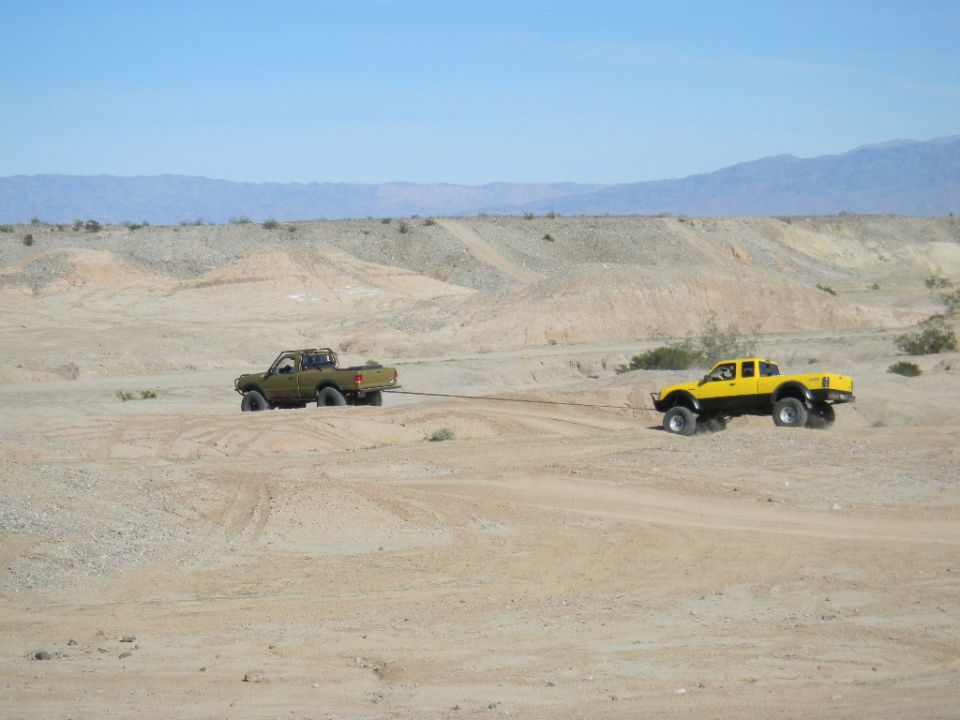 Yellow pickup truck towing a green pickup truck with a tow rope in a desert
