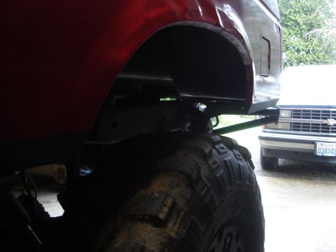1989 Ford Bronco - Low rear-quarter view under the back corner of a maroon lifted truck body, showing the empty rear wheel well, exposed frame rail and a hitc…