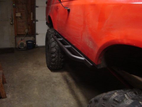 1989 Ford Bronco - The side of a red SUV is shown with large tires and custom side steps, parked indoors.