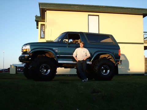 1996 Ford Bronco - A person stands beside a dark green Ford Bronco modified with oversized tires and lifted suspension.